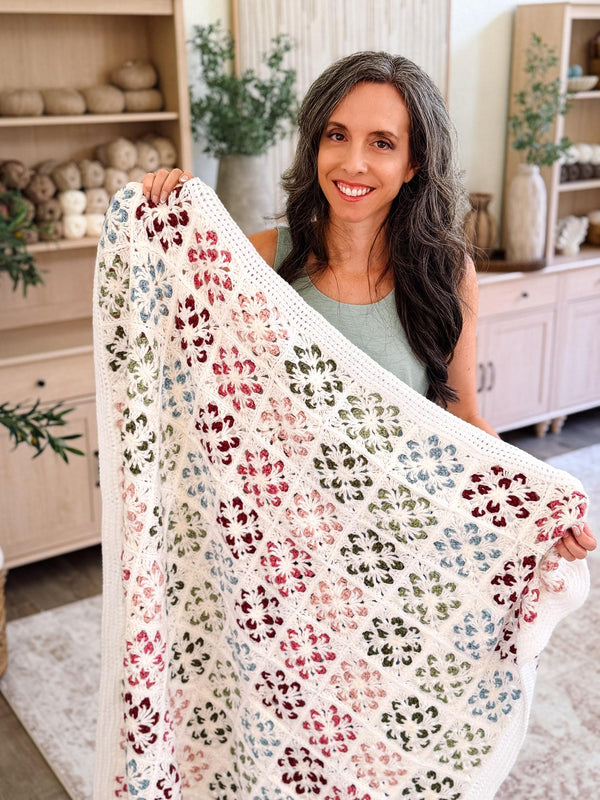 Blossom Granny Square Blanket crochet pattern: cream crochet blanket with red, pink, green and blue flower squares held up by a smiling woman in a yarn-filled room